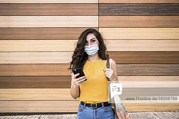 Junge Frau hält Smartphone und Netztasche  während sie an einer Holzwand steht