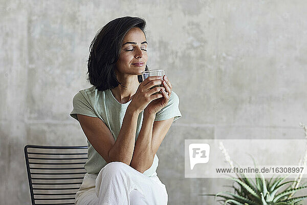 Businesswoman with eyes closed holding smoothie while sitting on chair against wall in office