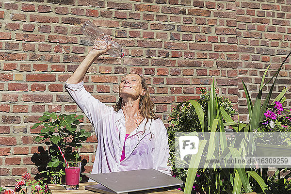 Geschäftsfrau erfrischt sich  indem sie Wasser aus einer Flasche auf dem Balkon gegen eine Backsteinmauer gießt  während einer Pause an einem sonnigen Tag