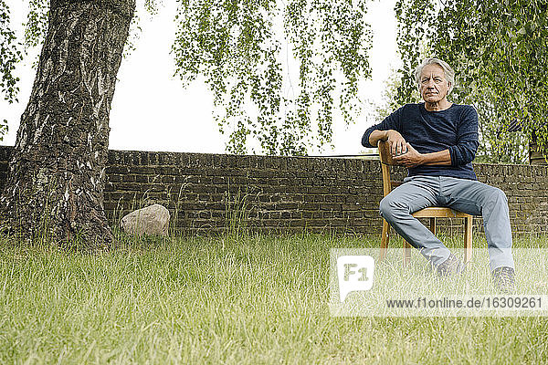 Contemplating man sitting on chair against brick wall in backyard
