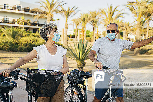 Älteres Paar mit Gesichtsmaske  das mit dem Fahrrad im Park spazieren geht