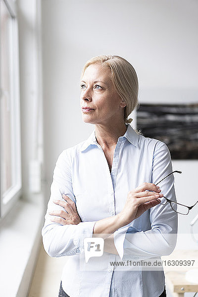 Thoughtful businesswoman with arms crossed holding eyeglasses while standing in home office