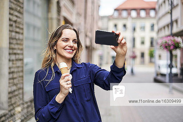 Lächelnde schöne Frau  die ein Selfie macht  während sie auf dem Fußweg ein Eis isst