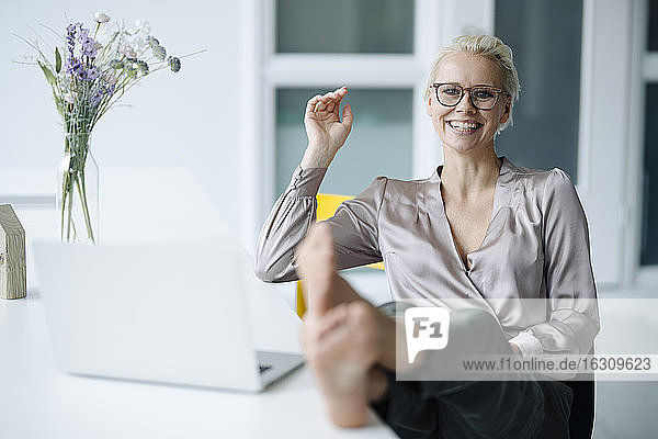 Lächelnde Geschäftsfrau mit den Füßen neben dem Laptop auf dem Schreibtisch beim Entspannen im Büro