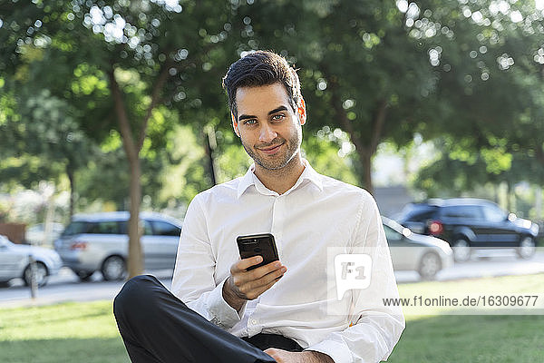 Confident businessman using smart phone while sitting against trees