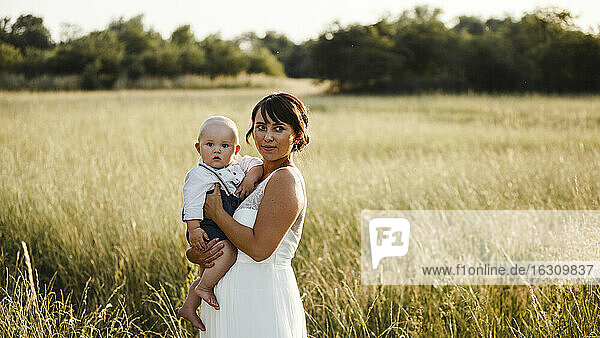 Bride carrying baby boy while standing at field during sunset