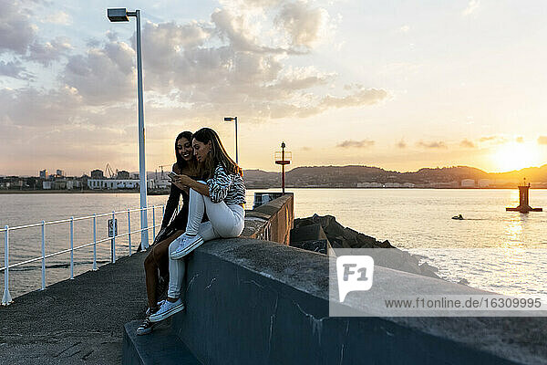 Friends using phone while sitting on retaining wall at promenade during sunset