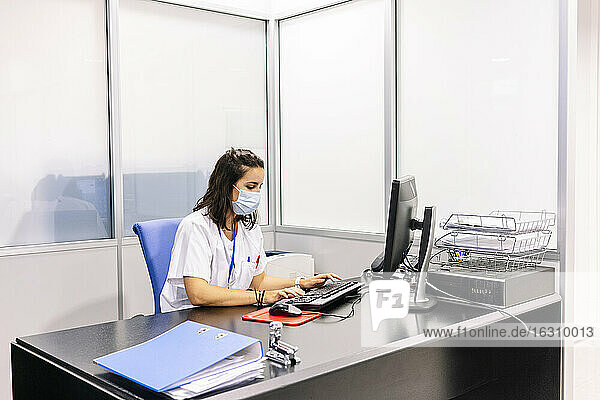 Weiblicher Arzt mit chirurgischer Maske  der einen Computer auf dem Schreibtisch im Büro eines Krankenhauses benutzt