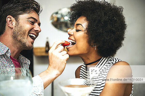 Close-up of cheerful man feeding strawberry to girlfriend in restaurant