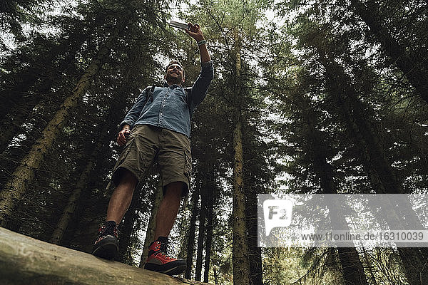 Mann  der mit seinem Smartphone fotografiert  während er gegen Bäume im Wald steht