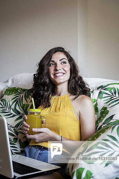 Thoughtful smiling woman holding juice while sitting with laptop on armchair at home