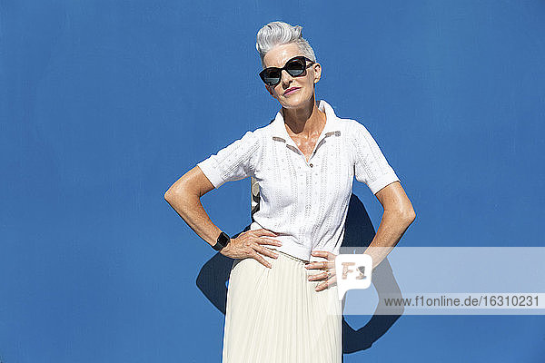Ältere Frau steht mit der Hand auf der Hüfte gegen eine blaue Wand an einem sonnigen Tag