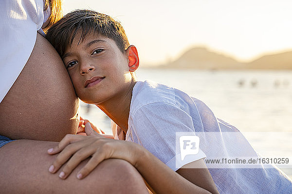 Sohn hält den Kopf auf dem Bauch der schwangeren Mutter  während er bei Sonnenuntergang am Strand sitzt