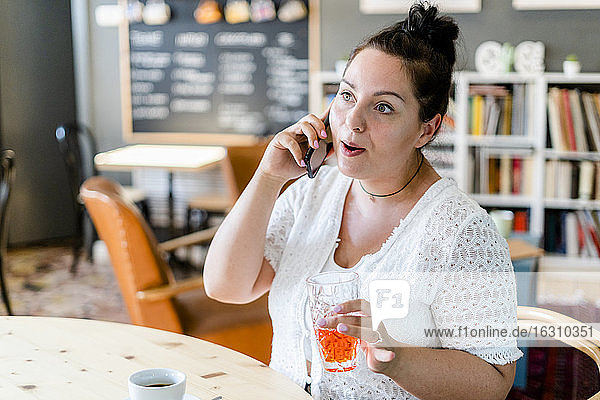Üppige Frau  die ein Getränk hält und über ein Mobiltelefon spricht  während sie im Restaurant sitzt