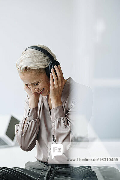Lächelnde Geschäftsfrau hört Musik über Kopfhörer  während sie im Büro an der Wand sitzt