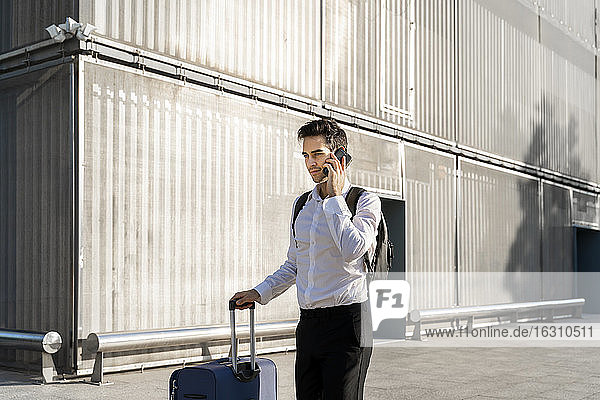Geschäftsmann mit Koffer  der über sein Smartphone spricht  während er auf dem Fußweg vor einem Gebäude steht
