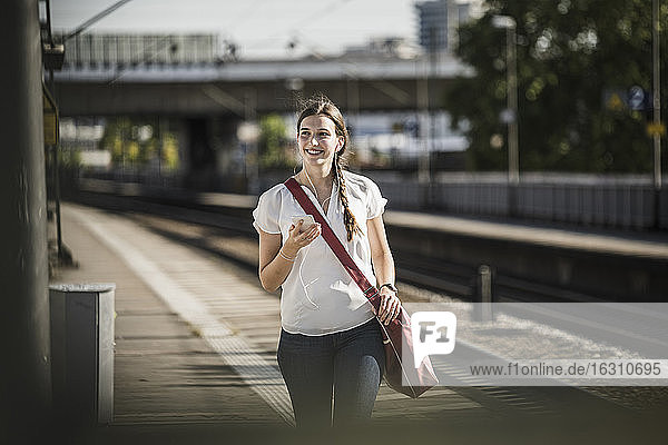 Lächelnde Frau hört Musik über ihr Mobiltelefon  während sie auf dem Bahnhof spazieren geht