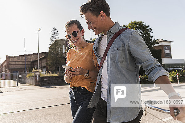 Lächelndes junges Paar  das auf sein Handy schaut  während es auf der Straße in der Stadt spazieren geht