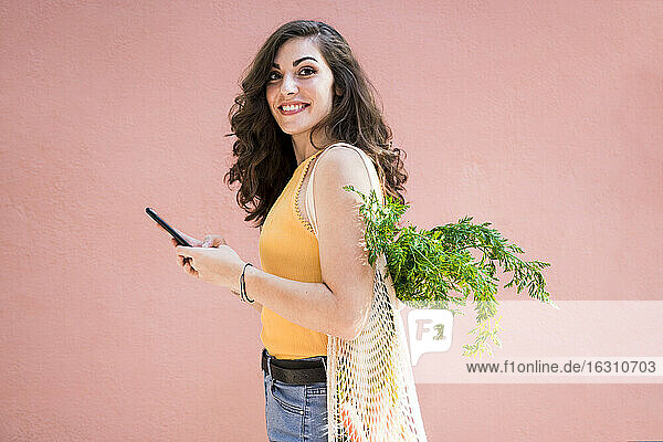 Lächelnde schöne junge Frau  die ihr Smartphone in der Hand hält  während sie mit einer wiederverwendbaren Netztasche vor einer rosa Wand steht