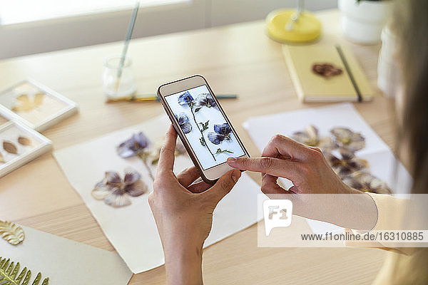 Hands of young woman photographing dry flowers and leaves on table at home
