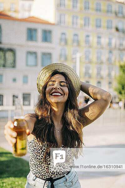 Cheerful young woman wearing hat holding beer bottle while standing in city