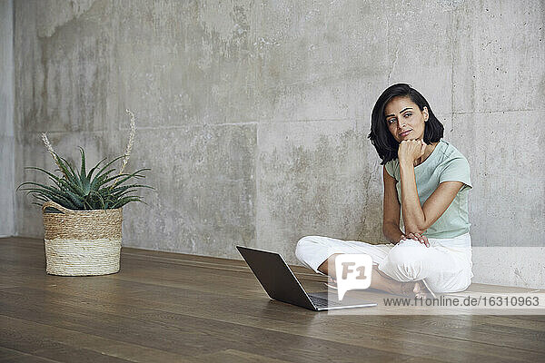 Nachdenkliche Unternehmerin sitzt mit Laptop auf Hartholzboden an der Wand im Büro