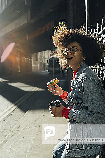 Afro woman holding donut and coffee while standing in city during sunny day