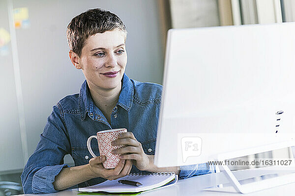 Lächelnde Geschäftsfrau am Schreibtisch im Büro mit Blick auf den Computerbildschirm