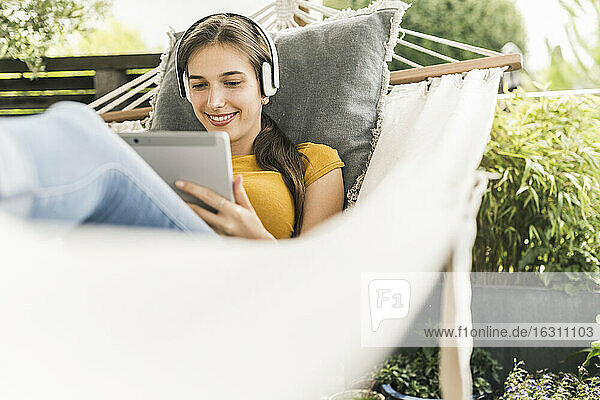 Lächelnde junge Frau mit Kopfhörern  die ein digitales Tablet benutzt  während sie sich in einer Hängematte im Garten entspannt