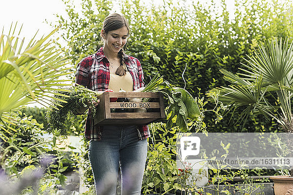 Smiling young woman carrying vegetables in crate while standing amidst plants at garden