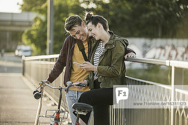 Smiling boyfriend and girlfriend looking at smart phone while standing with bicycle in city