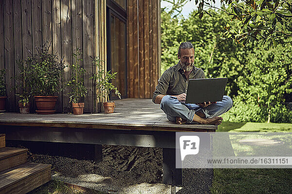 Älterer Mann  der einen Laptop benutzt  während er an einem kleinen Haus sitzt