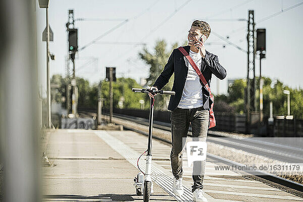 Lächelnder Mann  der mit einem Mobiltelefon spricht  während er mit einem Roller auf dem Bahnsteig spazieren geht
