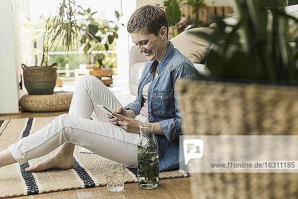 Lächelnde erwachsene Frau mit kurzen Haaren  die ein Smartphone benutzt  während sie zu Hause auf einem Teppich sitzt