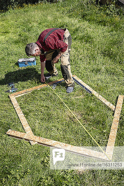 Zimmermann misst Spielhausbretter  die an einem sonnigen Tag auf den Rasen gelegt werden