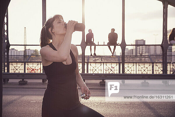 Frau trinkt Wasser aus einer Flasche  nachdem sie auf einer Brücke bei Sonnenuntergang in der Stadt trainiert hat