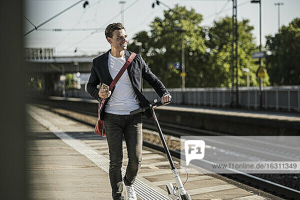 Smiling young man looking away while walking with push scooter on railroad station platform