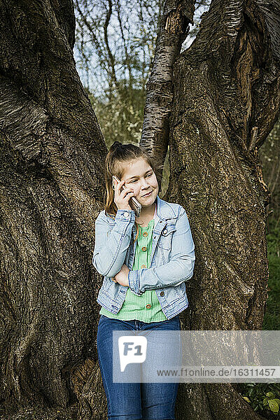 Lächelndes Mädchen  das mit seinem Smartphone spricht  während es gegen einen Baumstamm im Park steht