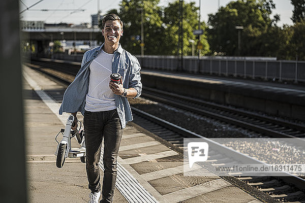 Lächelnder junger Mann mit Tretroller auf dem Bahnsteig