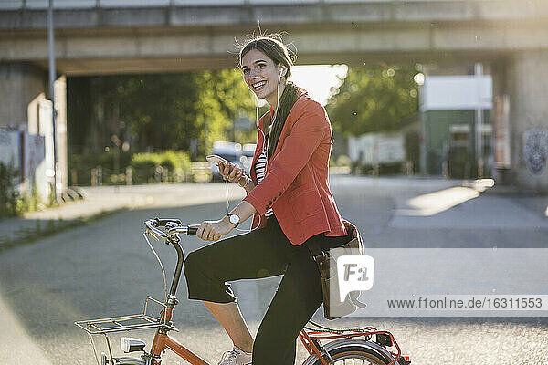 Happy young woman using smart phone while riding bicycle on street in city