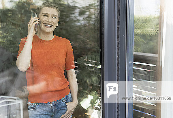 Fröhliche Frau  die zu Hause über ihr Smartphone spricht  gesehen durch ein Fenster