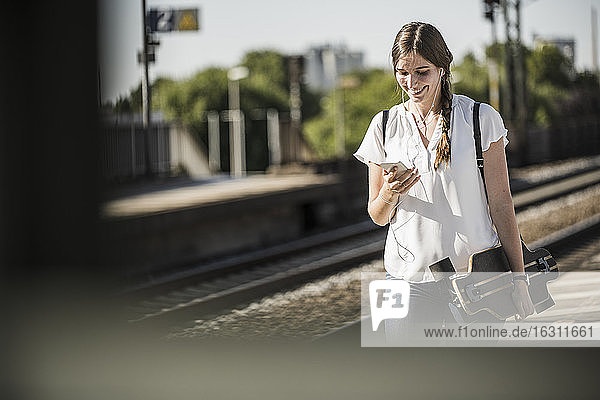 Smiling young woman with skateboard using smart phone at railroad station