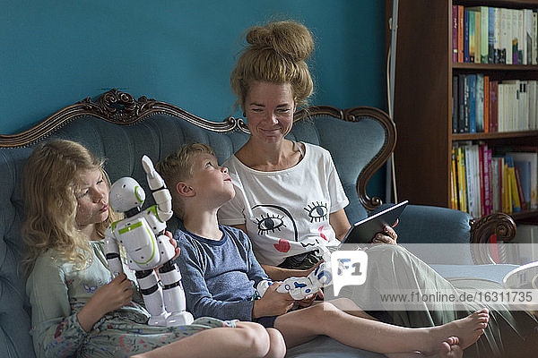Lächelnde Frau betrachtet Kinder  die mit Robotern spielen  die auf dem Sofa im Wohnzimmer zu Hause sitzen
