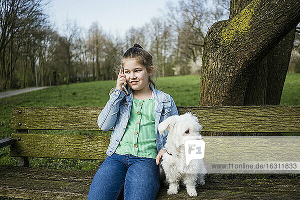 Lächelndes Mädchen mit Hund telefoniert auf einer Bank im Park