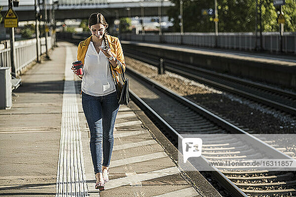 Lächelnde junge Frau  die ein Smartphone benutzt  während sie auf dem Bahnsteig spazieren geht
