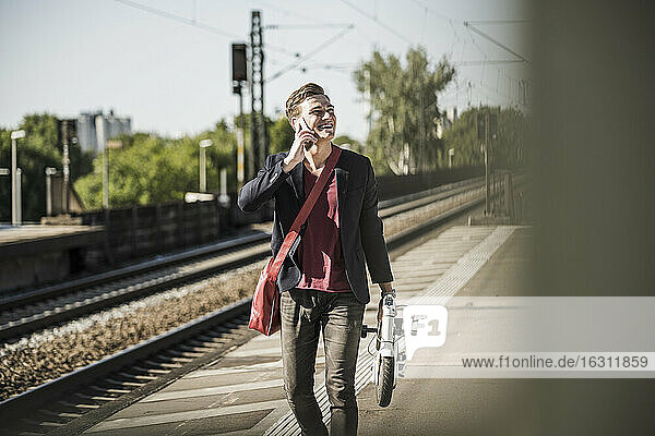 Mann mit Tretroller telefoniert am Bahnsteig mit Handy