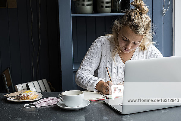 Woman sitting alone at a cafe table with a laptop computer  writing in note book  working remotely.
