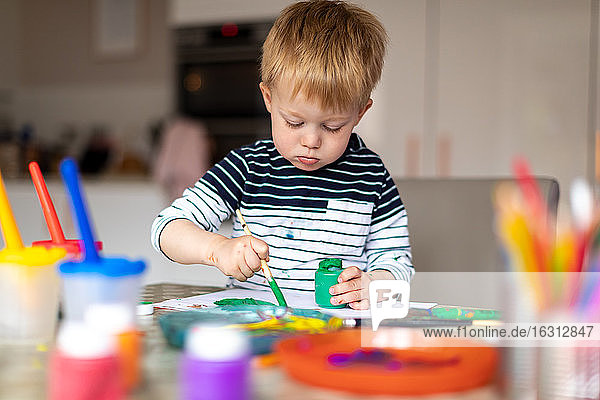 Three year old boy busy painting at home  with paint pots and brushes.