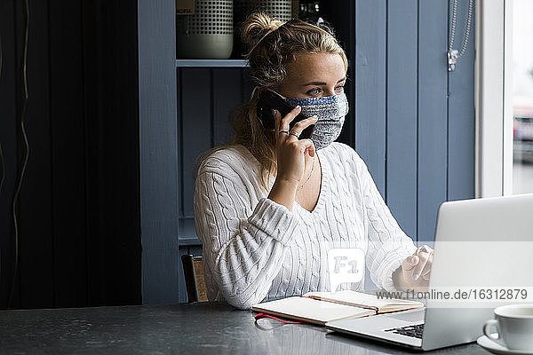 Frau mit Gesichtsmaske  die an einem Café-Tisch sitzt  Mobiltelefon und Laptop benutzt und aus der Ferne arbeitet.