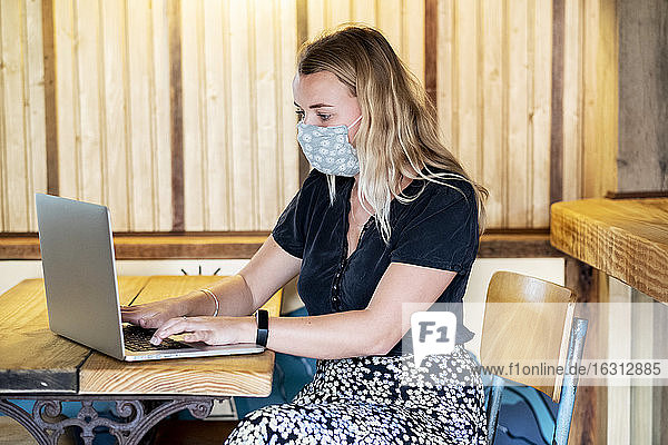 Young blond woman wearing blue face mask  sitting at table  using laptop computer.